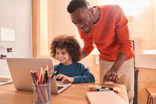 Easy task. Pleasant smart bearded man standing behind his curly son while helping him with homework. Father helping and support concept. Stock photo