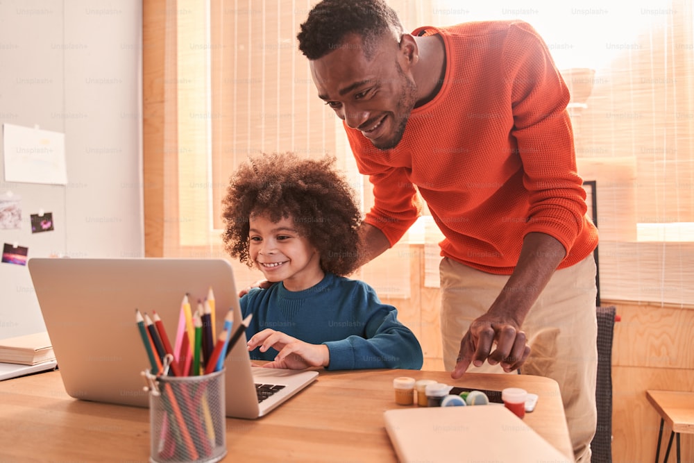 Easy task. Pleasant smart bearded man standing behind his curly son while helping him with homework. Father helping and support concept. Stock photo