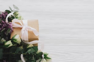 Happy mothers day and womens day concept. Stylish gift box with lilac and roses bouquet on white wooden background in soft light, space for text. Craft present with ribbon and spring flowers