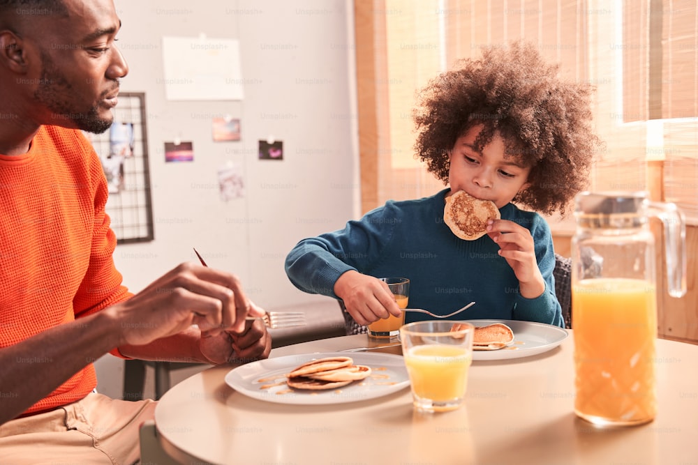 Male friendship. Father in casual clothes and his curly son eating pancakes and orange juice. Funny son holding pancake in his hands. People talking at the kitchen with friendly atmosphere