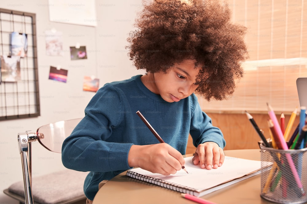 부엌에서 공부하는 동안 집에서 연필로 그림을 그리는 귀여운 미취학 아동 곱슬 소년의 초상화. 전염병 때문에 온라인 교육을 받는 어린 소년. 스톡 사진
