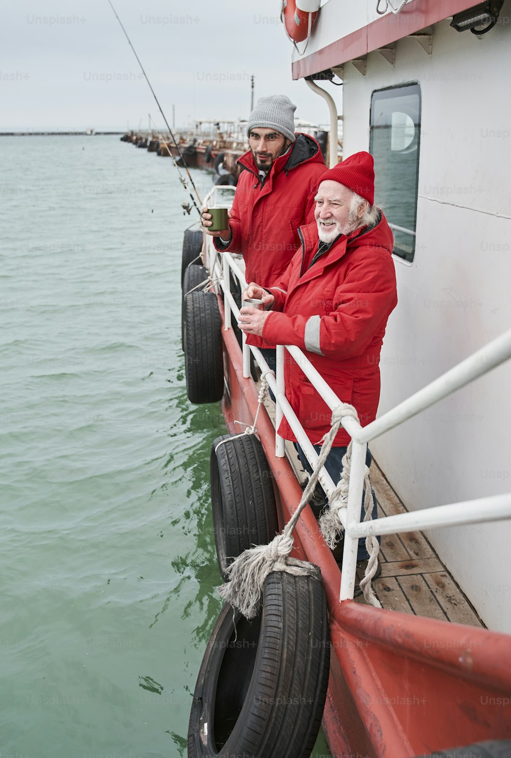 Professional fishing. Full length view of the senior bearded man telling something to his young colleague while drinking tea at the deck of the boat. Stock photo
