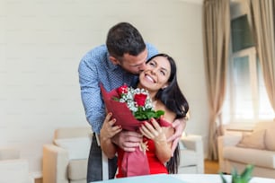 Affectionate young man giving his beautiful young wife a bouquet of red roses and kissing her on Valentines day , romantic happy couples sharing gift together on Valentines day