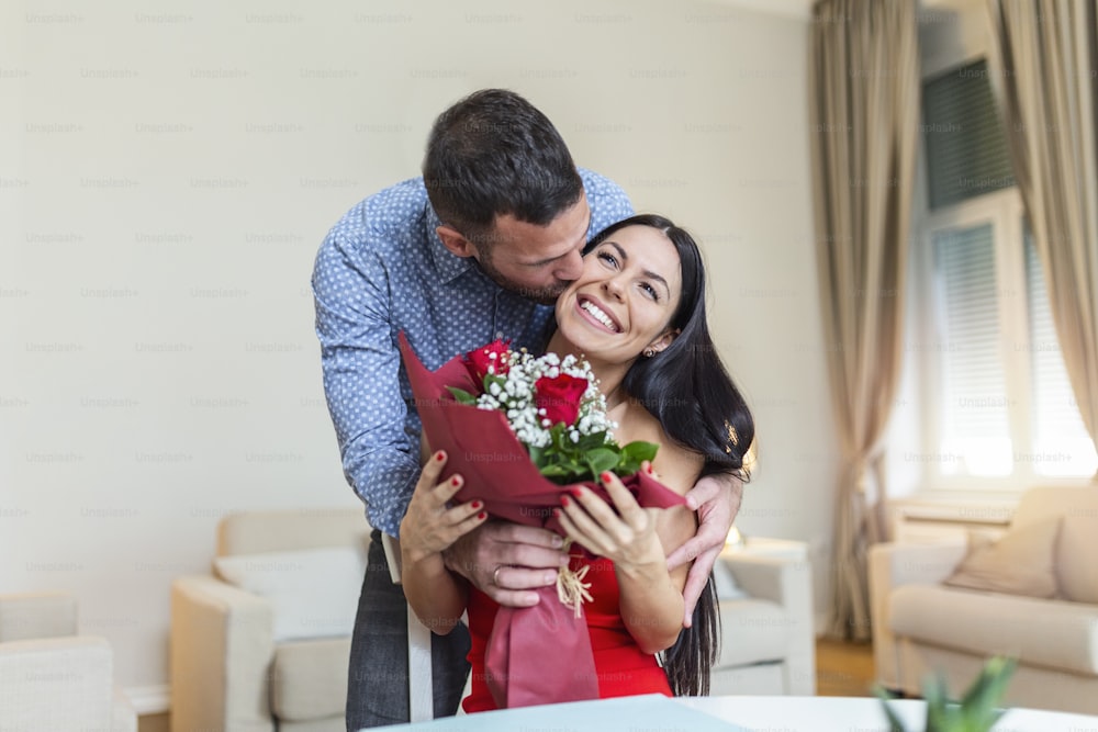 Affectionate young man giving his beautiful young wife a bouquet of red roses and kissing her on Valentines day , romantic happy couples sharing gift together on Valentines day
