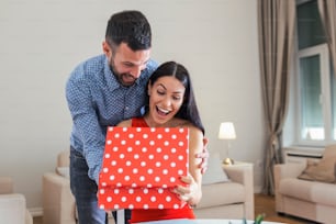 Excited young woman opening gift box receiving good unexpected present from husband at home, loving generous boyfriend making romantic surprise to attractive girlfriend on Valentines day occasion