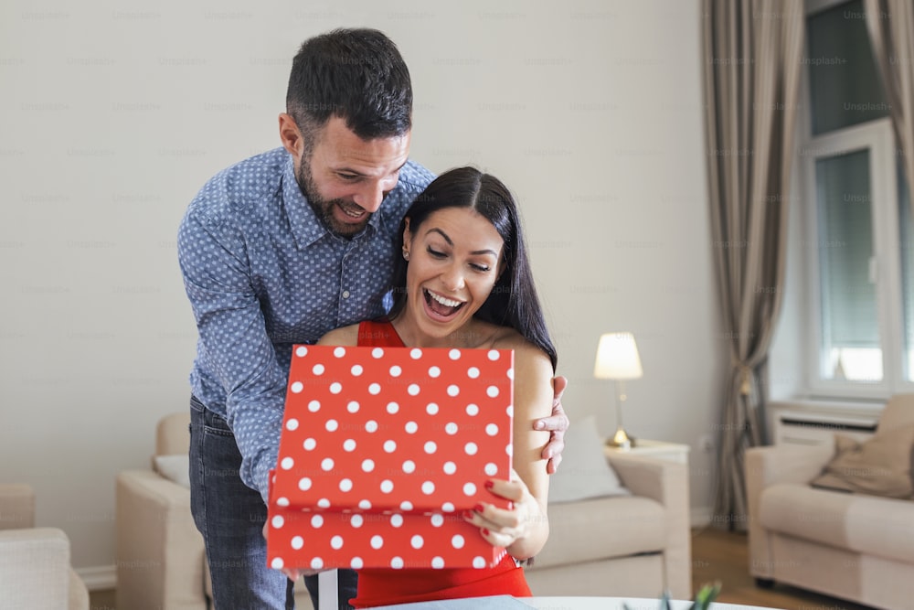 Excited young woman opening gift box receiving good unexpected present from husband at home, loving generous boyfriend making romantic surprise to attractive girlfriend on Valentines day occasion