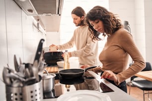 She is the center of his universe. Beautiful young couple cooking dinner in the kitchen at home. Man preparing toasts and his wife pushing button at the stove. Coolinary concept