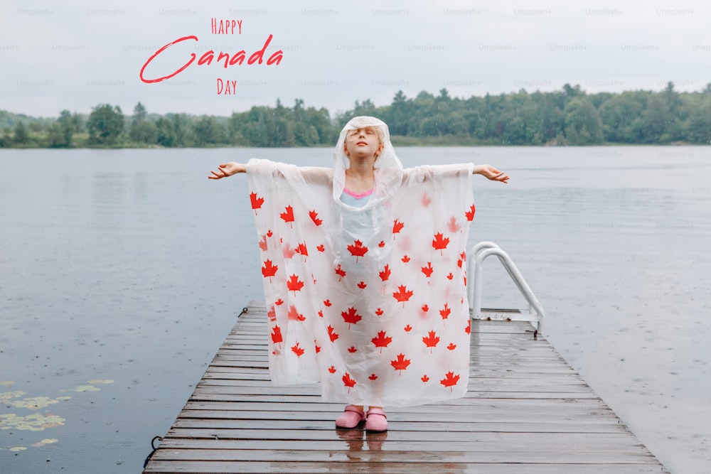 Feliz Día de Canadá. Tarjeta navideña con texto. Niña en poncho de lluvia con hojas de arce rojo de pie en el muelle de madera del lago. Niño levantando los brazos bajo la lluvia al aire libre. Conexión con la naturaleza.