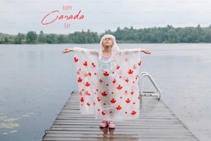 Happy Canada Day. Holiday card with text. Girl child in rain poncho with red maple leaves standing on wooden lake dock. Kid raising arms up under rain outdoor. Connection with nature.