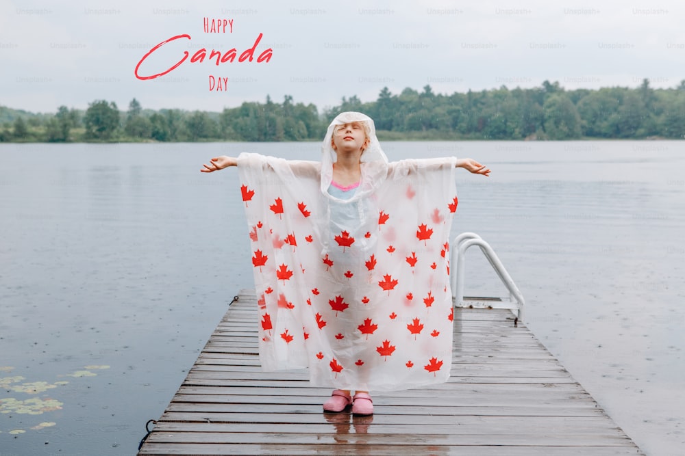Happy Canada Day. Holiday card with text. Girl child in rain poncho with red maple leaves standing on wooden lake dock. Kid raising arms up under rain outdoor. Connection with nature.