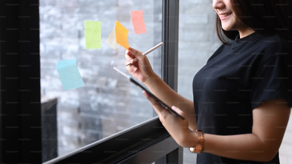 Confident businesswoman holding digital tablet and reading sticky notes on glass wall in office.