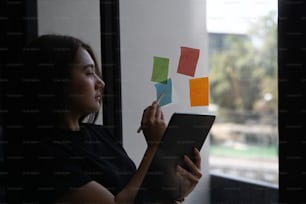 Businesswoman holding digital tablet and writing ideas on sticky notes in office.