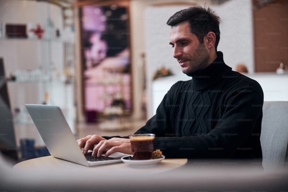 Hübscher, fröhlicher junger Mann macht Geschäfte online auf dem Laptop, während er sich bei heißem Kaffee entspannt