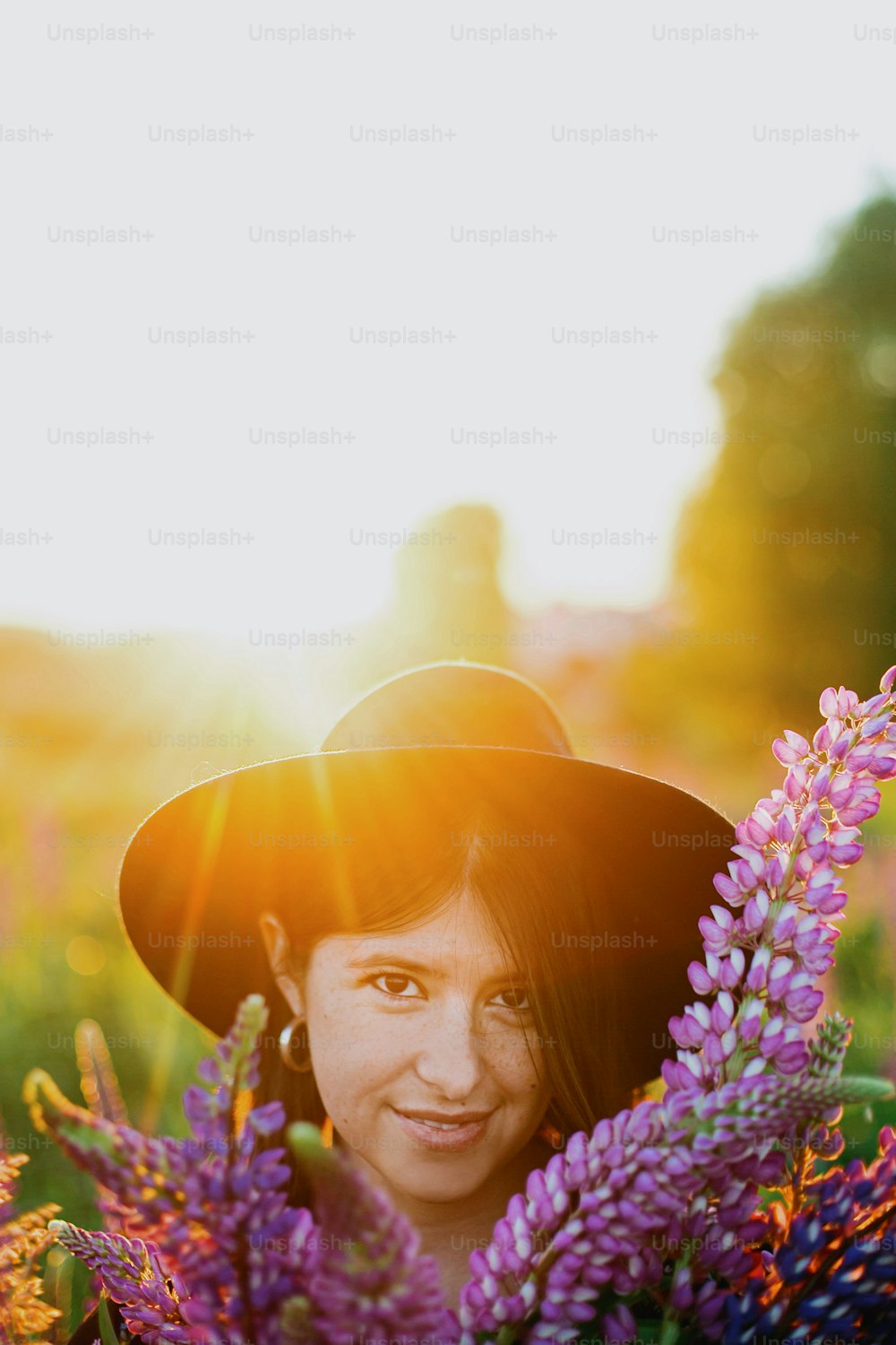 Beautiful woman relaxing in sunny lupine field, holding rustic basket with flowers. Tranquil atmospheric moment. Portrait of young female among purple wildflowers in countryside meadow at sunset