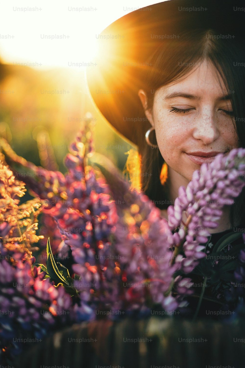 Portrait of beautiful woman holding lupine bouquet in sunset light in countryside field. Tranquil atmospheric moment. Young female with lupine flowers in warm sunshine in summer meadow