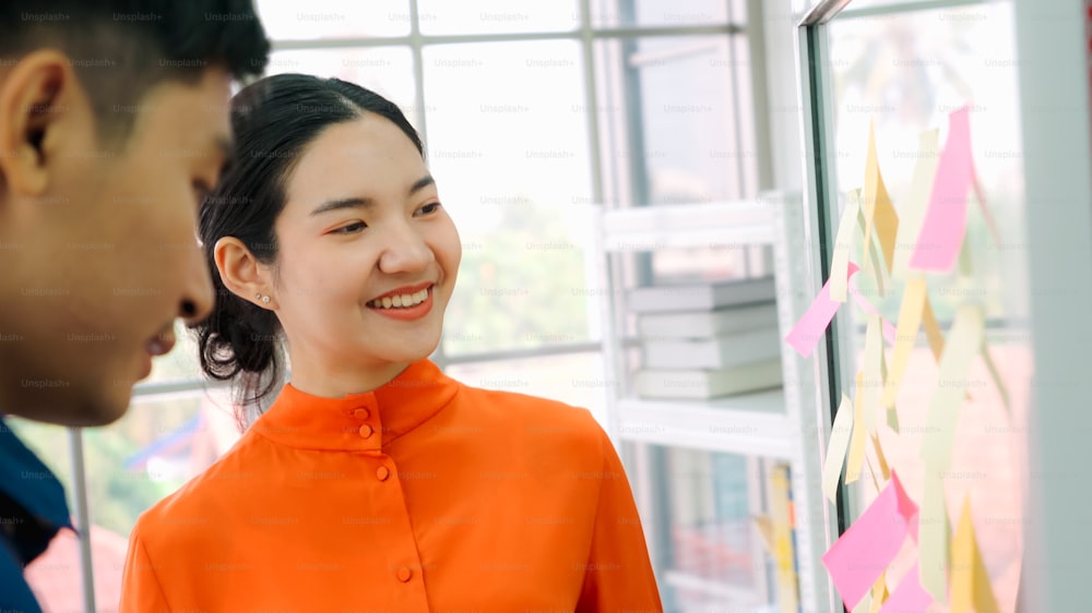 Business people work on project planning board in office and having conversation with coworker friend to analyze project development . They use sticky notes posted on glass wall to make it organized .