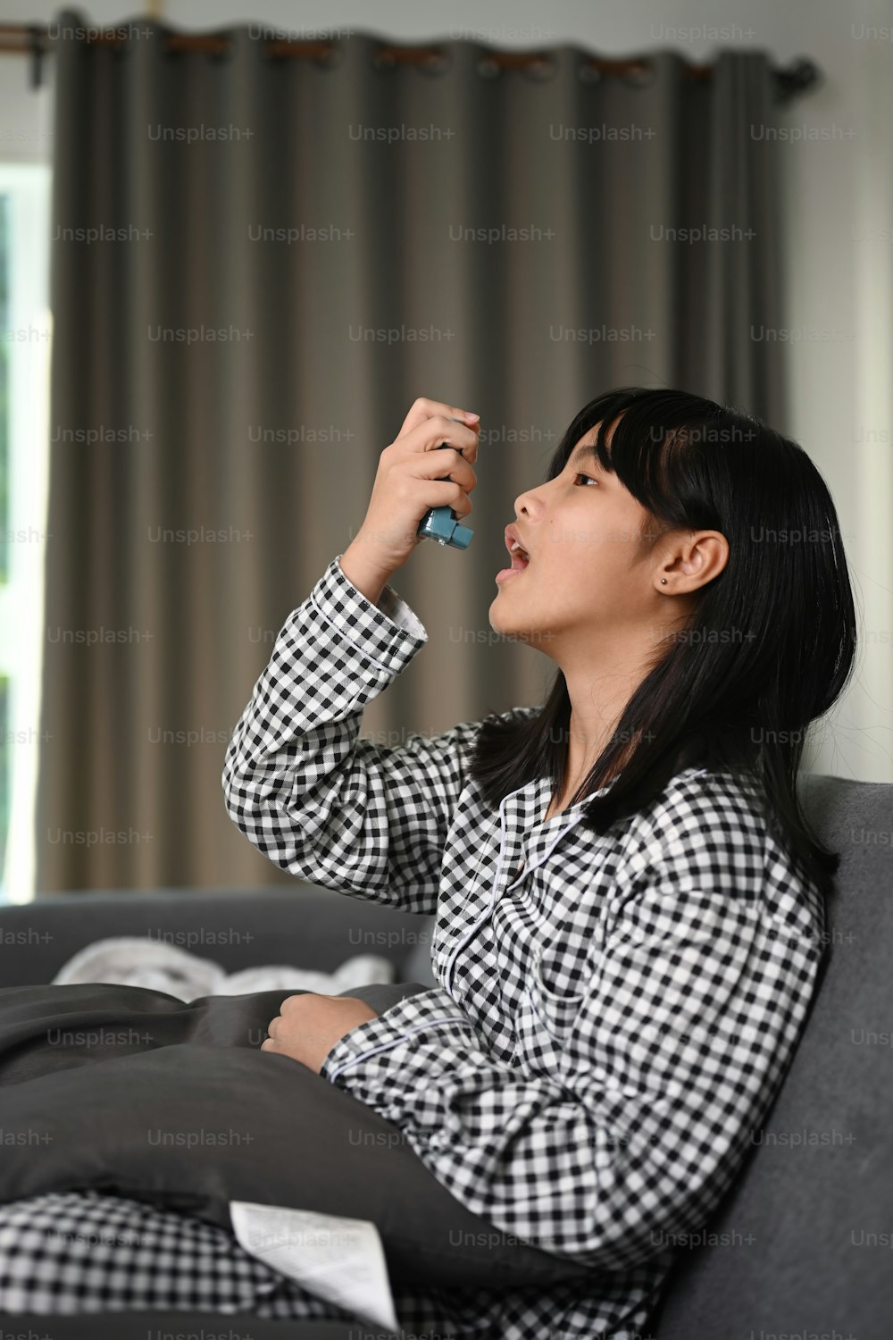 Image young girl using inhaler by herself for asthma while sitting on sofa at home.