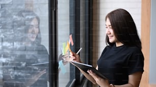 Smiling businesswoman holding digital tablet and reading sticky notes on glass wall in office.