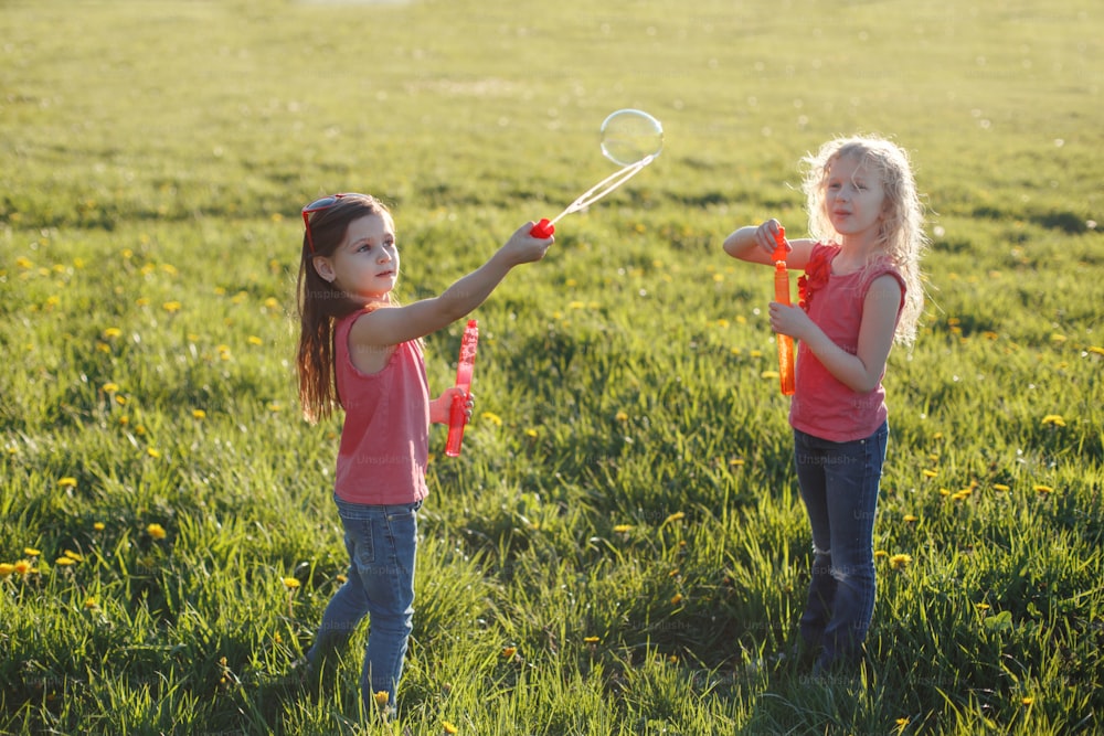 Happy Caucasian girls blowing soap bubbles in park on summer day. Kids having fun outdoor. Authentic happy childhood magic moment. Lifestyle seasonal activity for children.
