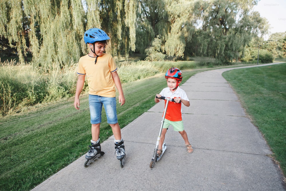 Caucasian boys brothers in helmets riding roller skates and scooter on road in park. Seasonal outdoor children activity sport. Healthy childhood lifestyle. Siblings friends relationships.
