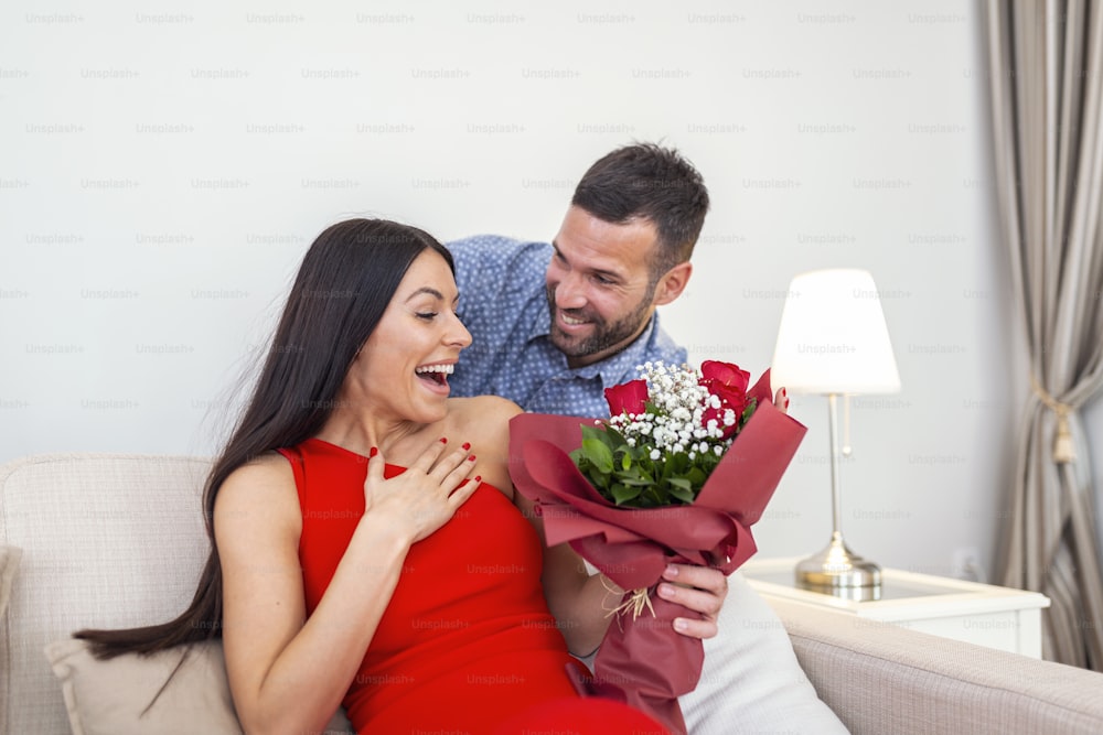 Excited young woman receiving unexpected Bouquet of red roses from husband at home, loving generous boyfriend making romantic surprise to attractive girlfriend on Valentines day occasion