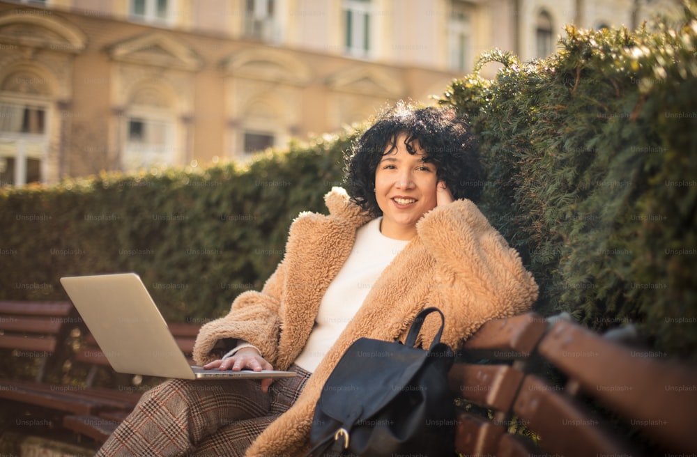 Lächelnde Frau, die auf einer Bank im Park sitzt und einen Laptop benutzt. Blick in die Kamera.