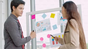 Business people work on project planning board in office and having conversation with coworker friend to analyze project development . They use sticky notes posted on glass wall to make it organized .