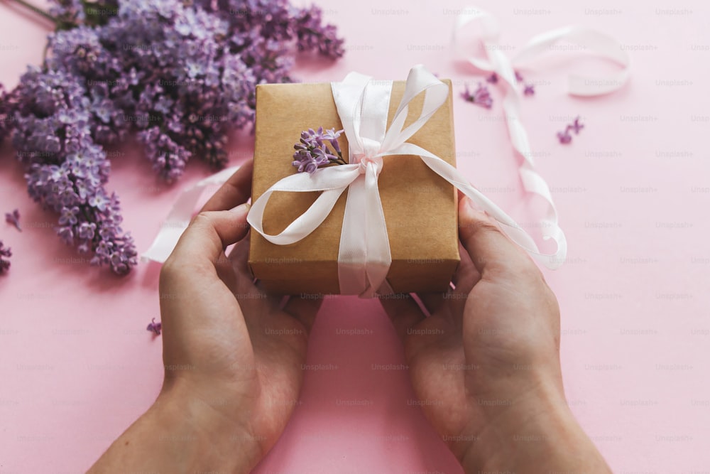Alles Gute zum Muttertag und zum Valentinstag. Hände halten Geschenkbox mit Band und lila Blumen auf rosa Papier. Lila lila Blumenstrauß mit Bastelgeschenkbox. Verschenken