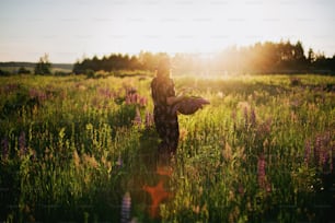 Beautiful stylish woman walking in sunny lupine field with wicker rustic basket and flowers. Tranquil atmospheric moment. Young female gathering wildflowers in summer countryside at sunset