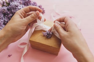 Happy mothers day and valentines day concept. Hands wrapping gift box with ribbon and lilac flowers on pink paper. Purple lilac flowers bouquet with craft present box