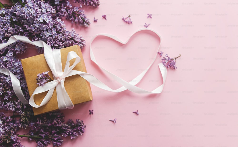 Fliederblumen, Geschenkbox und Herzband auf rosa Papier, flach gelegt mit Kopierraum. Stilvolle Blumengrußkarte. Alles Gute zum Valentinstag und Muttertagskonzept. Lila Fliederstrauß und Bastelgeschenk