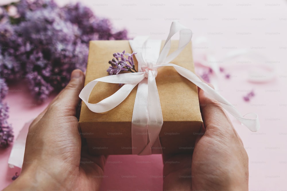 Hands holding gift box with ribbon and lilac flowers on pink paper. Happy mothers day and valentine's day congratulations concept. Purple lilac flowers bouquet with craft present box.