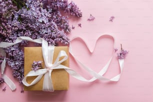 Lilac flowers, gift box and heart ribbon on pink paper, flat lay. Happy valentines day and mothers day concept. Purple lilac bouquet and craft present