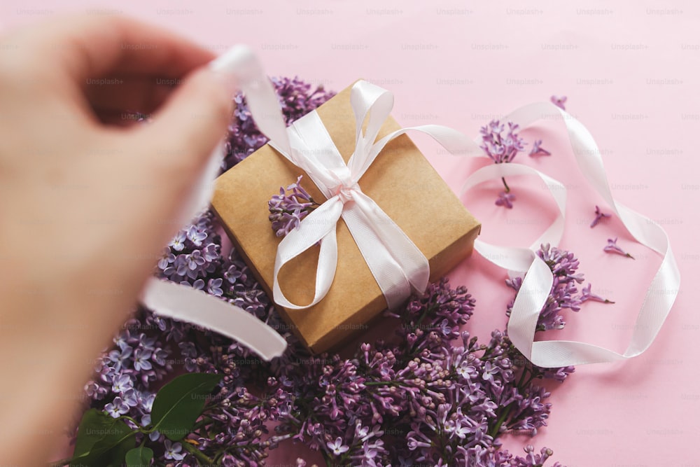 Hand opening gift box with ribbon and lilac flowers on pink paper, view above. Happy mothers day and valentine's day concept. Purple lilac flowers bouquet with craft present box