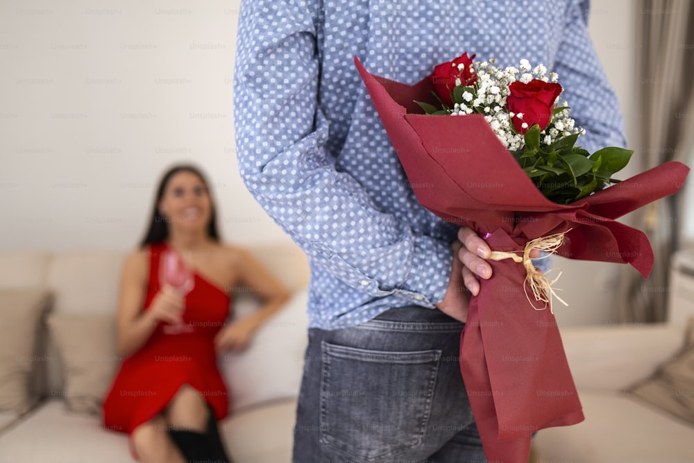 Happy woman looking at her boyfriend holding bouquet of roses behind his back. Handsome man holding flowers behind his back to surprise his girlfriend, romantic happy couples on Valentines day