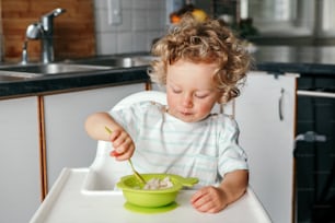 Simpatico adorabile ragazzo riccio caucasico seduto nel seggiolone che mangia purea di cereali con cucchiaio. Alimentazione sana per bambini, bambini. Bambino che mangia in modo indipendente. Momento candido e autentico.
