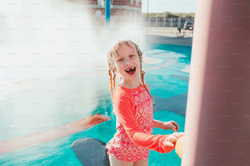 Süßes entzückendes lachendes kaukasisches lustiges Mädchen, das am Sommertag auf dem Splash-Pad-Spielplatz im Freien spielt. Saisonale Wassersport-Freizeitaktivität für Kinder.