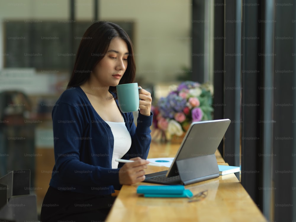 Seitenansicht einer Studentin, die Kaffee trinkt, während sie Hausaufgaben mit digitalem Tablet im Café macht