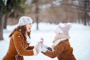 Madre e hija modernas sonrientes en sombreros de punto y abrigos de piel de oveja con manoplas en un sombrero de punto y abrigo de piel de oveja jugando afuera en el parque de la ciudad en invierno.