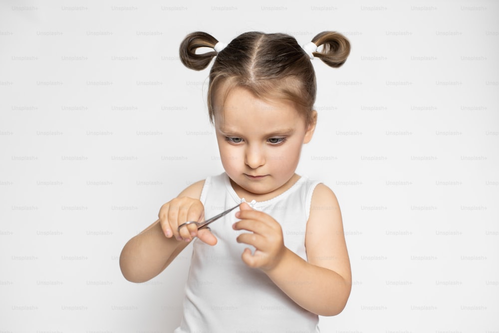 Close up portrait of little cute concentrated 3 years old kid girl, wearing white top and having ponytails hair style, cutting trimming her finger nails. Isolated on white.