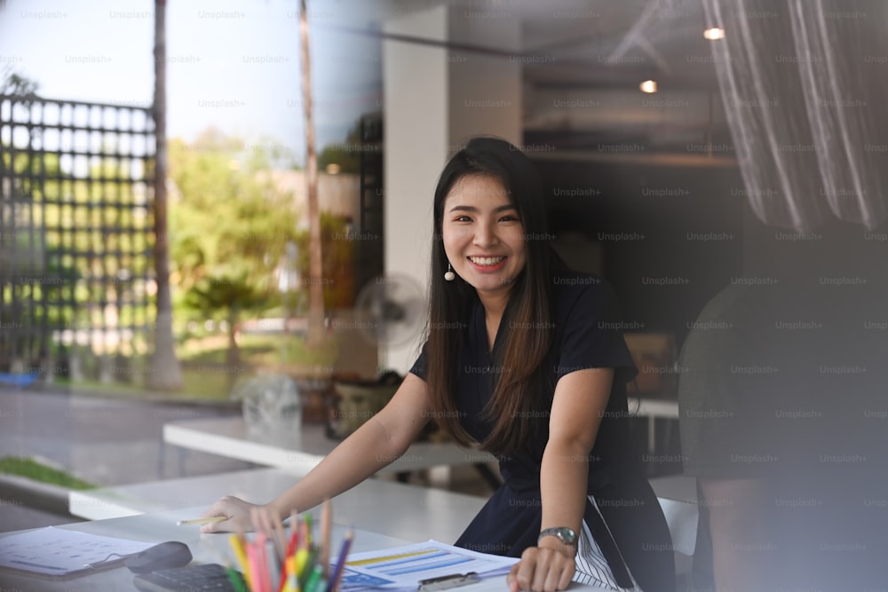 Smiling young female designer sitting at creative workplace and smiling to camera seen through glass at office.