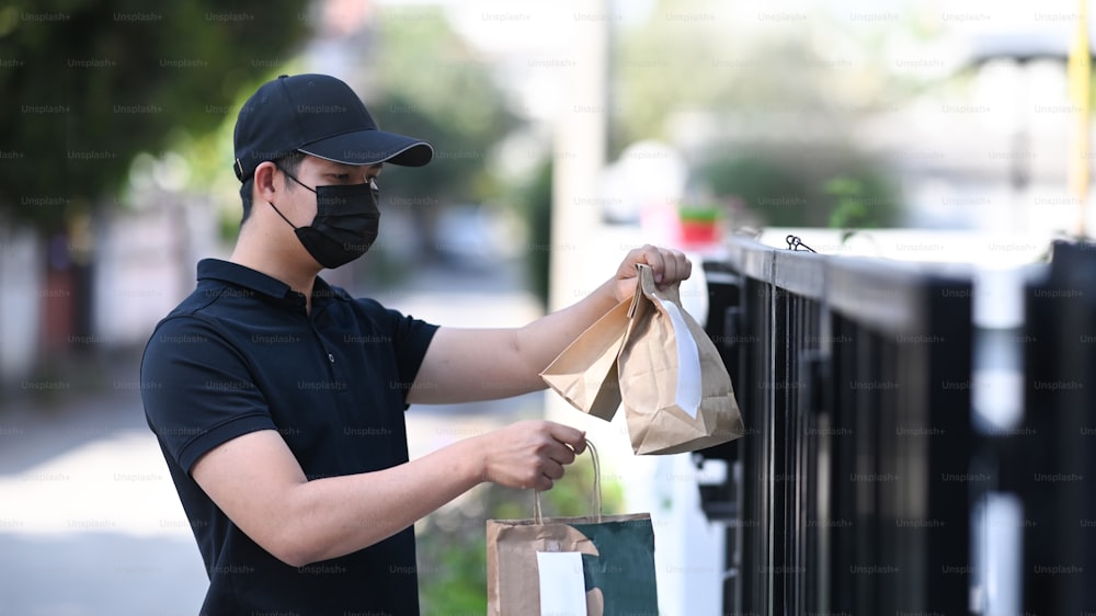 防護マスクを着けた若いアジア人の配達員が、食べ物の入った紙袋を持ち、家のドアで客を待っている。