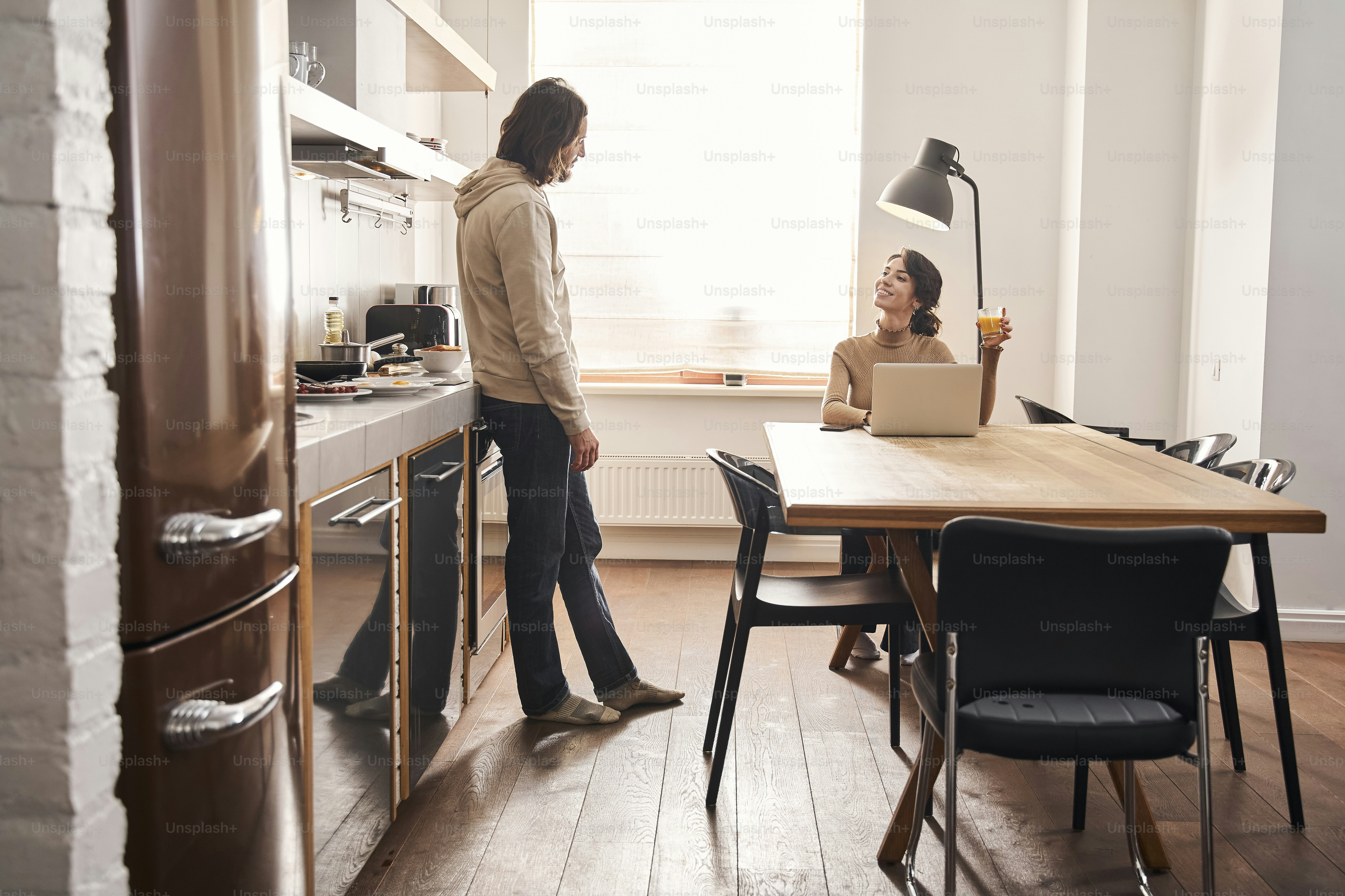 Heureux couple marié discutant pendant qu’une femme lit les nouvelles des médias sociaux sur un ordinateur portable dans une cuisine blanche fraîche. Les jeunes mariés profitent d’une journée de détente. Banque d’images