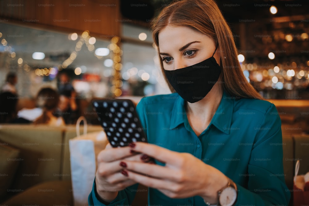 Beautiful young adult woman with protective face mask sitting in a nice restaurant and using her smart phone. She is alone and serious. Coronavirus pandemic and social distancing problems.