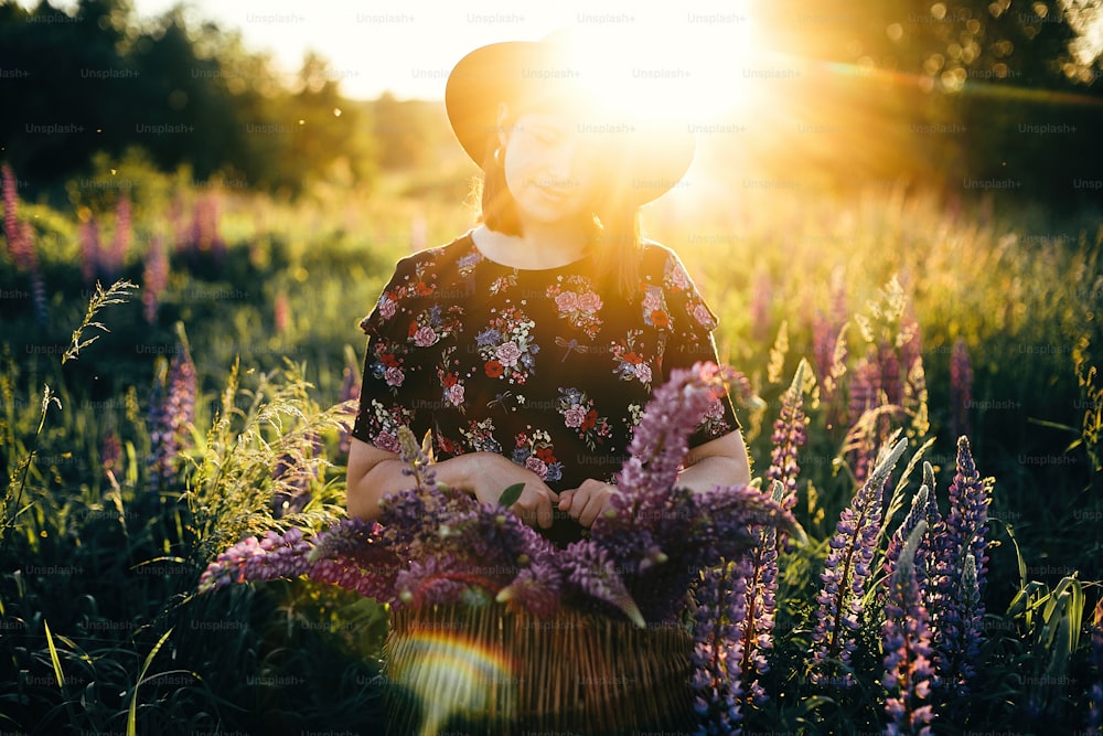 Beautiful stylish woman relaxing in sunny lupine field and holding rustic basket with flowers. Tranquil atmospheric moment. Young female gathering wildflowers in countryside meadow at sunset