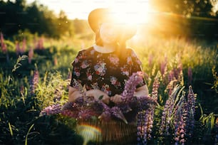 Beautiful stylish woman relaxing in sunny lupine field and holding rustic basket with flowers. Tranquil atmospheric moment. Young female gathering wildflowers in countryside meadow at sunset