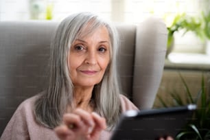 태블릿을 사용하여 집 소파에 실내에 앉아 있는 행복한 노인 여성의 초상화.