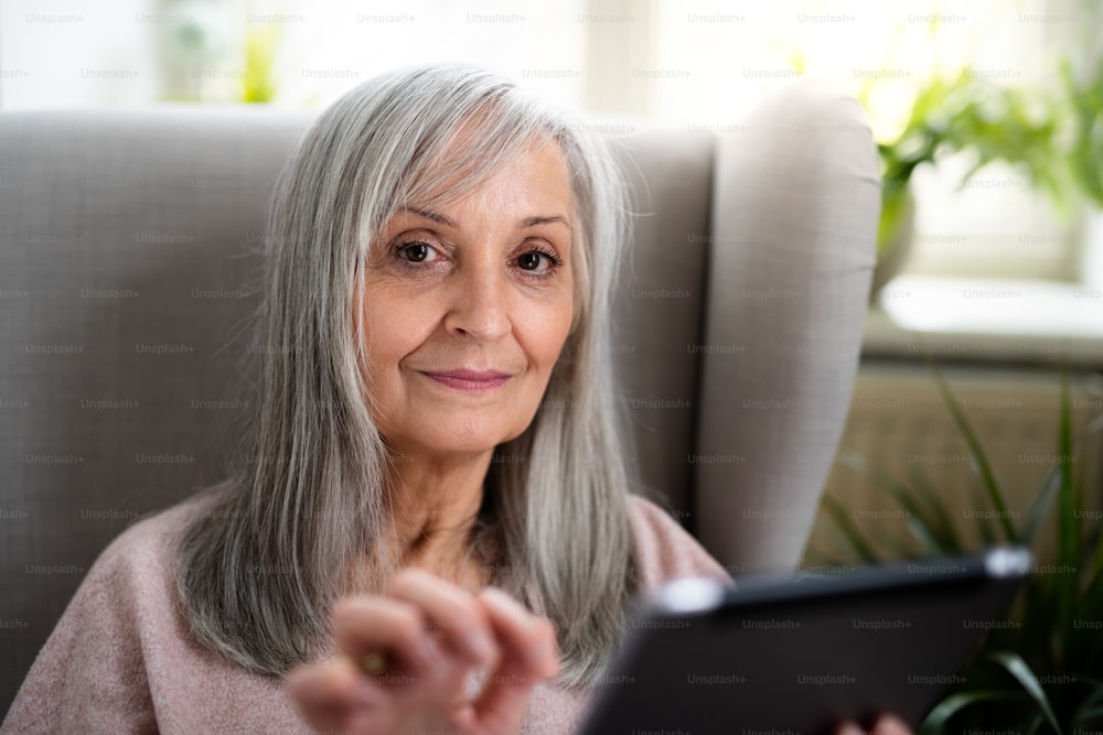 Ritratto di donna anziana felice seduta all'interno sul divano di casa, utilizzando il tablet.