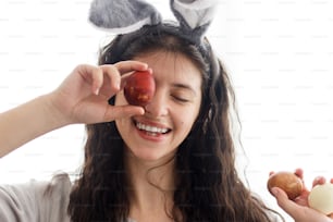 Happy young woman in bunny ears and linen dress holding easter eggs at eyes and smiling in white light. Natural dyed easter eggs in hands, funny moments. Happy Easter!