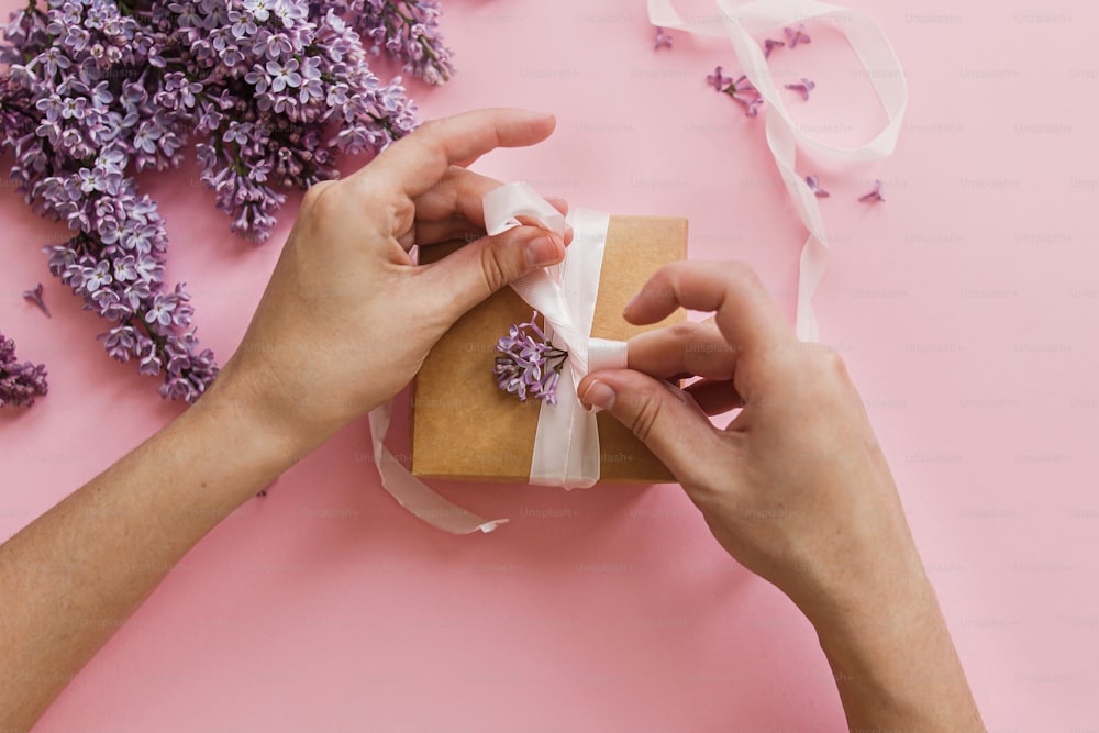 Hands wrapping gift box with ribbon and lilac flowers on pink paper, top view. Happy mothers day and valentines day concept. Purple lilac flowers bouquet with craft present box